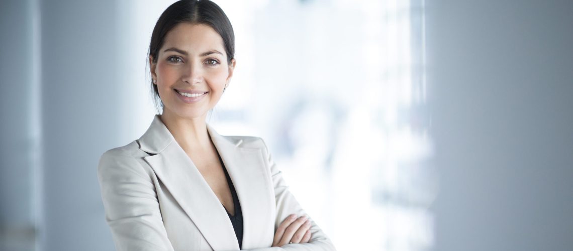 Closeup portrait of smiling beautiful middle-aged business woman wearing jacket and standing in light office hall with her arms crossed